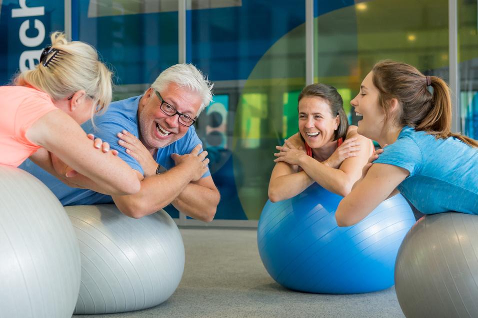 Vier Personen sitzen lachend auf Gymnastikbällen in einem hellen Fitnessraum und interagieren spielerisch miteinander.
